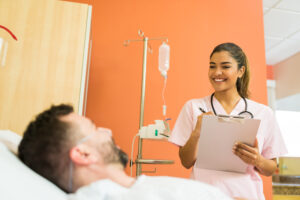 Profissional de saúde sorridente conversa com paciente em leito hospitalar, segurando uma prancheta para anotações, simbolizando atendimento acolhedor e gestão humanizada.