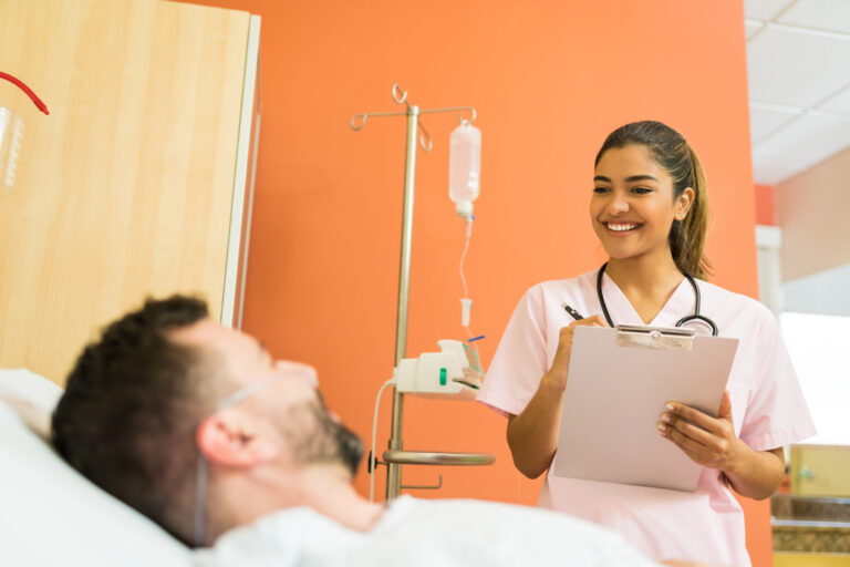 Profissional de saúde sorridente conversa com paciente em leito hospitalar, segurando uma prancheta para anotações, simbolizando atendimento acolhedor e gestão humanizada.